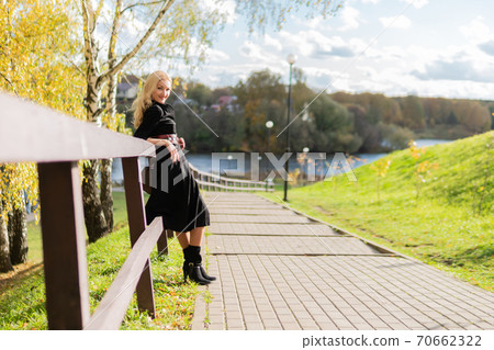 Happy stands and leans on the railing, an adult in black clothes, in the fall against a background of trees, blue sky. Happy stands and leans on the railing, an adult in black clothes, in the fall against a background of trees, blue sky. 70662322