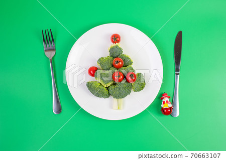 Christmas tree of organic vegetables on white plate; Broccolis and tomatoes with fork and knife on green background. Merry Xmas and happy New Year party, New You, Healthy food and vegetarian concept Christmas tree of organic vegetables on white plate; Broccolis and tomatoes with fork and knife on green background. Merry Xmas and happy New Year party, New You, Healthy food and vegetarian concept 70663107