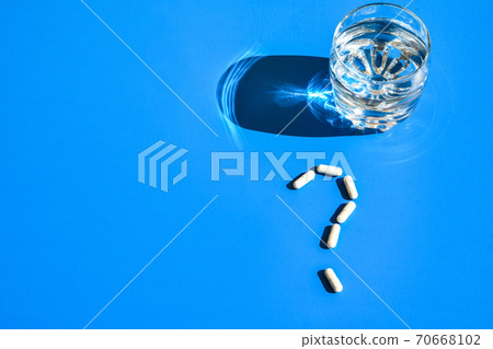 Glass of water and pills in shape of question mark on blue background. Hard light and shadows. Vitamins and prebiotics, probiotics. Dose Glass of water and pills in shape of question mark on blue background. Hard light and shadows. Vitamins and prebiotics, probiotics. Dose 70668102