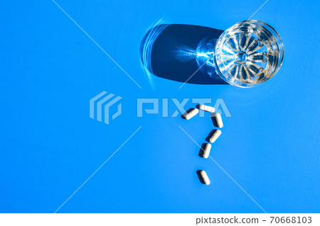 Glass of water and pills in shape of question mark on blue background. Hard light and shadows. Vitamins and prebiotics, probiotics. Dose Glass of water and pills in shape of question mark on blue background. Hard light and shadows. Vitamins and prebiotics, probiotics. Dose 70668103