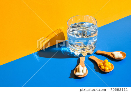 Glass of water and pills on wooden spoon on blue and yellow background with hard light and shadows. Modern isometric creative minimalism concept. Vitamins and prebiotics, probiotics. Dose Glass of water and pills on wooden spoon on blue and yellow background with hard light and shadows. Modern isometric creative minimalism concept. Vitamins and prebiotics, probiotics. Dose 70668111