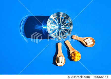 Glass of water and pills on wooden spoon on blue background with hard light and shadows. Creative. Vitamins and prebiotics, probiotics. Dose 70668331
