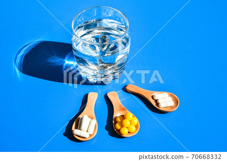 Glass of water and pills on wooden spoon on blue background with hard light and shadows. Creative. Vitamins and prebiotics, probiotics. Dose Glass of water and pills on wooden spoon on blue background with hard light and shadows. Creative. Vitamins and prebiotics, probiotics. Dose 70668332