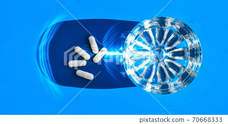 Glass of water and white pills on blue background with hard light and shadows. Creative. White tablets and drink on blue background. Vitamins and prebiotics Glass of water and white pills on blue background with hard light and shadows. Creative. White tablets and drink on blue background. Vitamins and prebiotics 70668333