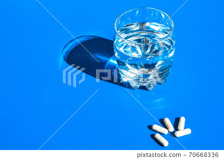 Glass of water and white pills on blue background with hard light and shadows. Creative. White tablets and drink on blue background. Vitamins and prebiotics Glass of water and white pills on blue background with hard light and shadows. Creative. White tablets and drink on blue background. Vitamins and prebiotics 70668336