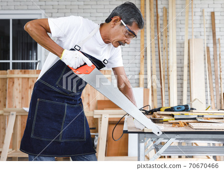 Senior carpenter sawing wood in carpentry workshop. Concept senior people doing hobby after retirement. Senior carpenter sawing wood in carpentry workshop. Concept senior people doing hobby after retirement. 70670466