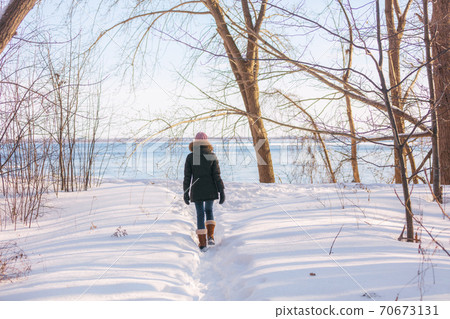 Winter hike woman walking in snow forest nature park by the river. Cold weather outerwear girl wearing warm down coat jacket and wool hat and boots Winter hike woman walking in snow forest nature park by the river. Cold weather outerwear girl wearing warm down coat jacket and wool hat and boots 70673131