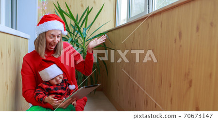 christmas online congratulations: a woman with a girl in red Santa hats congratulating family or friends with a tablet. the mother and daughter celebrates the new year in isolation. Winter holidays 70673147