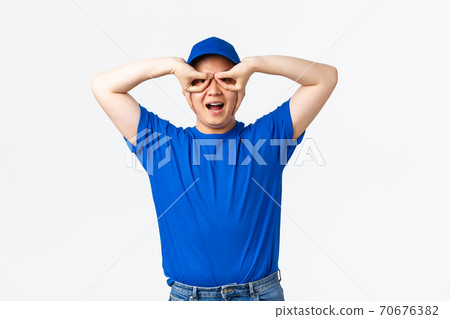 Carefree smiling asian courier in blue uniform, mimicking superhero mask, showing hand glasses. Funny delivery guy standing over white background, ensure parcels are safe and sound 70676382
