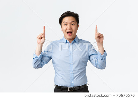 Business, finance and people concept. Excited and amazed asian businessman in blue shirt making announcement, pointing fingers up and look camera, telling big news, white background 70676576