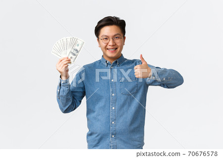 Smiling satisfied asian man in glasses and braces trying work e-commerce, freelancer got payment, showing thumbs-up and money, show-off his first paycheck, standing white background 70676578