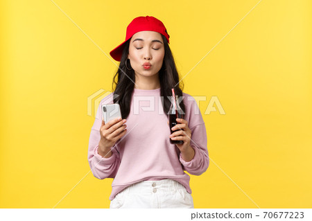 People emotions, drinks and summer leisure concept. Silly and cute asian girl in red cap, close eyes and fold lips to send air kiss, using smartphone and drinking soda from bottle, yellow background 70677223