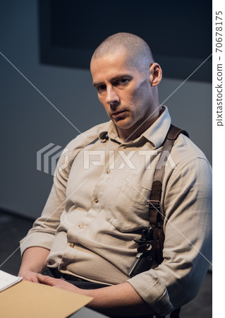 Bald investigator tired after questioning, portrait. A brutal man with a gun holster in a dark room with dramatic light. 70678175