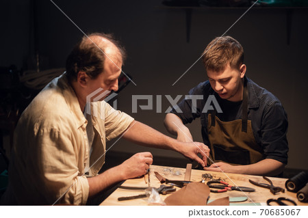 an elderly shoemaker and his apprentice create shoes by hand in their workshop with various tools. 70685067