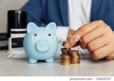 Blue piggy bank, stamp and man's fingers with coins on wooden desk in office. Save money and management financial concept 70689597