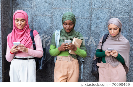 Three Modern Muslim Women Using Cellphones Standing Over Gray Wall 70690469