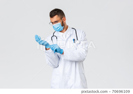 Covid-19, healthcare workers, pandemic, preventing virus concept. Young doctor in white scrubs and medical mask put on rubber gloves to observe patient in clinic, working during coronavirus outbreak Covid-19, healthcare workers, pandemic, preventing virus concept. Young doctor in white scrubs and medical mask put on rubber gloves to observe patient in clinic, working during coronavirus outbreak 70696109