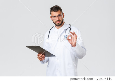 Healthcare workers, medical insurance, clinic lab and covid-19 concept. Handsome serious doctor in white coat, glasses and clipboard, show okay sign, assure tests alright, results are good Healthcare workers, medical insurance, clinic lab and covid-19 concept. Handsome serious doctor in white coat, glasses and clipboard, show okay sign, assure tests alright, results are good 70696110