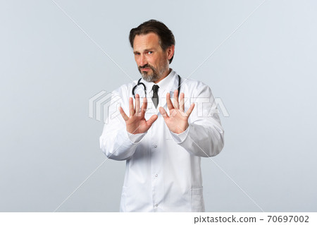 Covid-19, coronavirus outbreak, healthcare workers and pandemic concept. Reluctant and displeased male doctor in white coat saying no, show refusal gesture, tell to stop or stay away Covid-19, coronavirus outbreak, healthcare workers and pandemic concept. Reluctant and displeased male doctor in white coat saying no, show refusal gesture, tell to stop or stay away 70697002