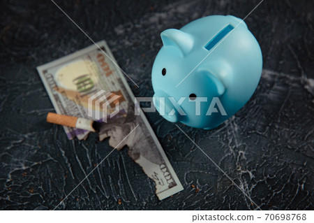 Blue piggy bank, dollar and cigarette on dark table. Expensive habit Blue piggy bank, dollar and cigarette on dark table. Expensive habit 70698768
