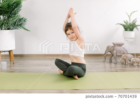 Healthy child girl, doing exercise in sportive top and leggings, practicing yoga at studio indoors. sitting in lotus pose on yoga mat with arms raised up, smiling to camera Healthy child girl, doing exercise in sportive top and leggings, practicing yoga at studio indoors. sitting in lotus pose on yoga mat with arms raised up, smiling to camera 70701439