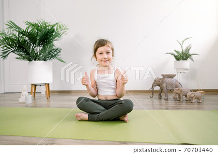 Happy smiling little Caucasian girl in sports suit practices yoga at home or fitness hall, sitting in lotus position on green yoga mat and showing her thumbs up Happy smiling little Caucasian girl in sports suit practices yoga at home or fitness hall, sitting in lotus position on green yoga mat and showing her thumbs up 70701440
