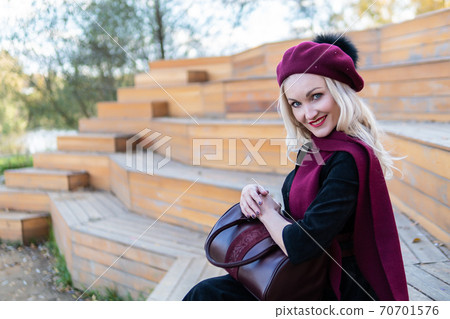 A smiling woman sits on a summer theater bench, made of wood in a burgundy coat and biret, an adult looks at the camera, in the fall against a background of blue clouds. 70701576