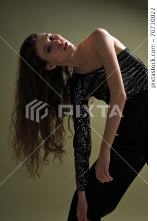 A beautiful young tall girl with long black hair poses in a shiny blouse and black culottes on a green background. Buty and fashion photo shoot. 70710022