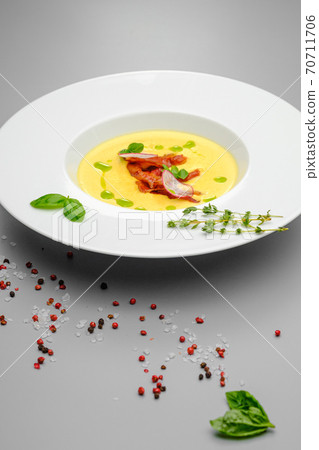 Corn cream soup with smoked meat in a white plate decorated with basil, on a gray background. The first course on the restaurant menu. 70711706