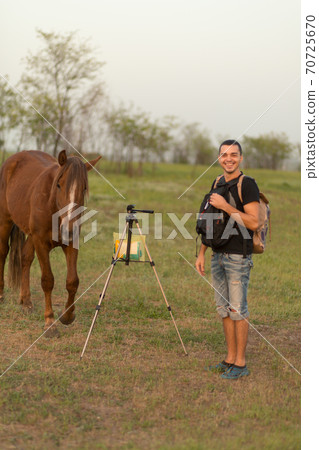 A guy with a tripod for a photo video shooting in a meadow with a horse. The horse is interested A guy with a tripod for a photo video shooting in a meadow with a horse. The horse is interested 70725670