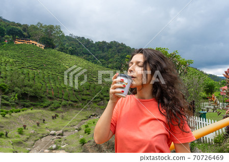 Brunette tourist girl enjoys tea from a craft cup against the background of a green tea valley. 70726469