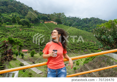 Brunette tourist girl enjoys tea from a craft cup against the background of a green tea valley. 70726470
