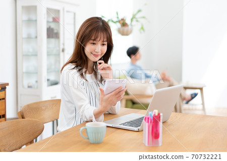 A wife smiling at a calculator at a dining table and a husband relaxing on a sofa 70732221