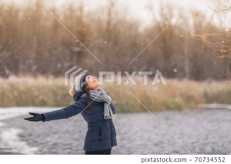 Winter happy girl enjoying snowing snowfall snowflakes falling. Asian lady with open arms wearing cold weather scarf, hat, gloves warm jacket walking outdoor nature woods background 70734552