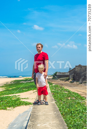 Father son walk along narrow concrete path, sea blue clear sky green grass background. Symbol as dad leads child into future adulthood. Happy childhood, fathers day, daddy influence on boy worldview Father son walk along narrow concrete path, sea blue clear sky green grass background. Symbol as dad leads child into future adulthood. Happy childhood, fathers day, daddy influence on boy worldview 70735289