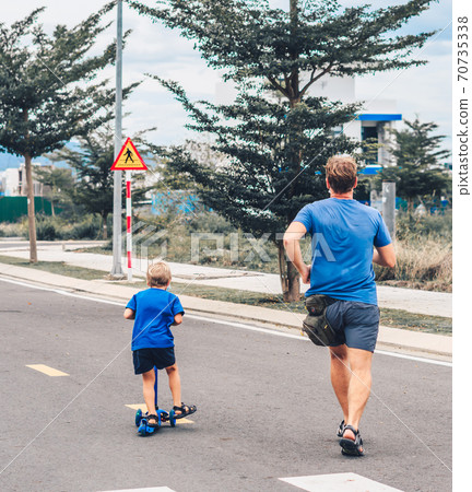 Coronavirus COVID 19 Sport active outdoor family life. Back view Father running son riding scooter movement, sidewalk background, having workout enjoying together. Concept run from problems Coronavirus COVID 19 Sport active outdoor family life. Back view Father running son riding scooter movement, sidewalk background, having workout enjoying together. Concept run from problems 70735338