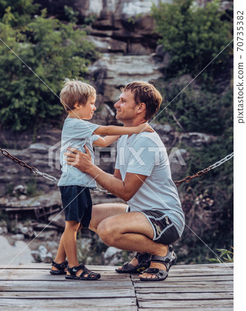 Father hugs son sitting squatting, look each other, smile. Happy childhood, fathers day, mountain, chain background as symbol of strong bond between dad and child. Father influence on son worldview Father hugs son sitting squatting, look each other, smile. Happy childhood, fathers day, mountain, chain background as symbol of strong bond between dad and child. Father influence on son worldview 70735382