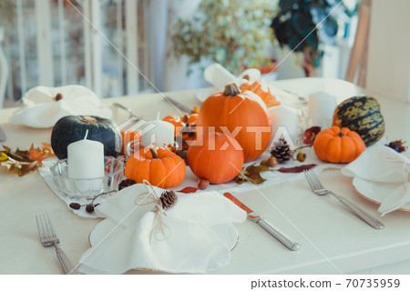 Festive table setting for Thanksgiving family home dinner. Fall composition with decorative pumpkins, nuts, cones, fallen leaves, and candles in light interior. Natural autumn decor. Selective focus Festive table setting for Thanksgiving family home dinner. Fall composition with decorative pumpkins, nuts, cones, fallen leaves, and candles in light interior. Natural autumn decor. Selective focus 70735959