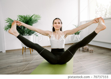 Front view of young woman yoga fitness coach, wearing sport top and leggins, stretching legs, sitting on green mat with outstretched legs in the air, in cozy yoga studio. Training for stretching 70737333