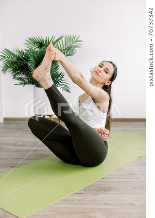 Pretty Caucasian girl in sport clothes performing stretching yoga exercise, sitting on green yoga mat, in modern training yoga hall. Yoga and healthy lifestyle concept. Pretty Caucasian girl in sport clothes performing stretching yoga exercise, sitting on green yoga mat, in modern training yoga hall. Yoga and healthy lifestyle concept. 70737342