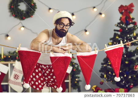 Funny Elf or Santa in underpants standing by line of drying red hats in his decorated house Funny Elf or Santa in underpants standing by line of drying red hats in his decorated house 70737863