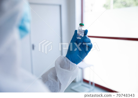 Rear view of health worker wearing protective clothes holding a vial bottle with Covid-19 text Rear view of health worker wearing protective clothes holding a vial bottle with Covid-19 text 70748343
