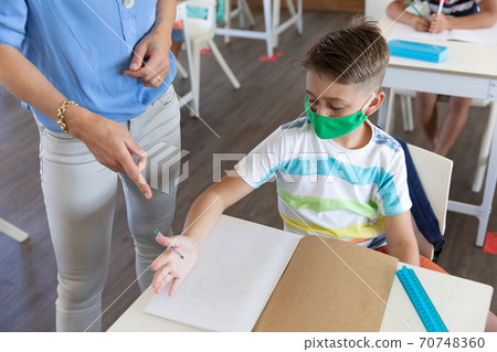 Mid section of female teacher wearing face mask teaching a boy in class at school 70748360