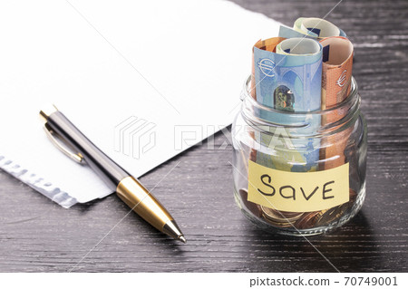 Euro bills and coins in a glass jar with a plate with a blank sheet of paper and a pen on a black wooden table with a sunny reflection on a glossy surface. Savings concept Euro bills and coins in a glass jar with a plate with a blank sheet of paper and a pen on a black wooden table with a sunny reflection on a glossy surface. Savings concept 70749001