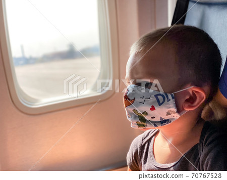 A caucasian child with light brown hair for about two years sits in a children's fabric protective mask with a drawn on board an airplane near an illuminator due to the spread of Covid-19 in the A caucasian child with light brown hair for about two years sits in a children's fabric protective mask with a drawn on board an airplane near an illuminator due to the spread of Covid-19 in the 70767528