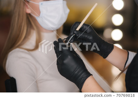 Close up of brow master hands in black gloves hold brush with henna. Background of customer in medical mask 70769818