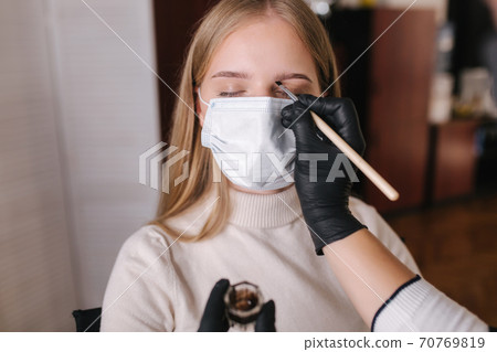Brow master in black protective mask applies henna on eyebrows for beautiful blond hair woman. Customer in medical mask. First person view 70769819