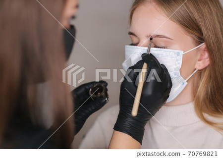 Brow master in black protective mask applies henna on eyebrows for beautiful blond hair woman. Customer in medical mask Brow master in black protective mask applies henna on eyebrows for beautiful blond hair woman. Customer in medical mask 70769821