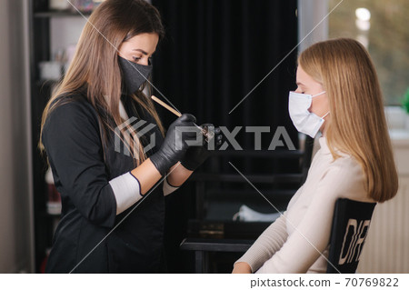 Brow master in black protective mask applies henna on eyebrows for beautiful blond hair woman. Customer in medical mask 70769822
