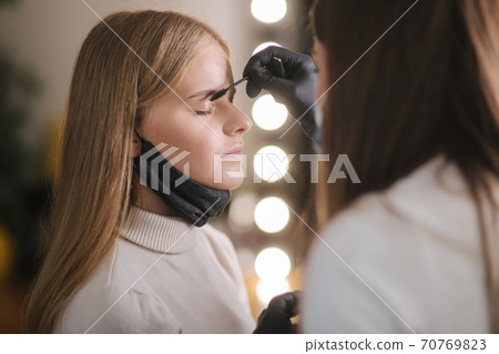 Brow and makeup master in protective mask gives shape to pull out with forceps previously painted with henna eyebrows in a beauty salon. Beautiful blond hair model in medical mask Brow and makeup master in protective mask gives shape to pull out with forceps previously painted with henna eyebrows in a beauty salon. Beautiful blond hair model in medical mask 70769823